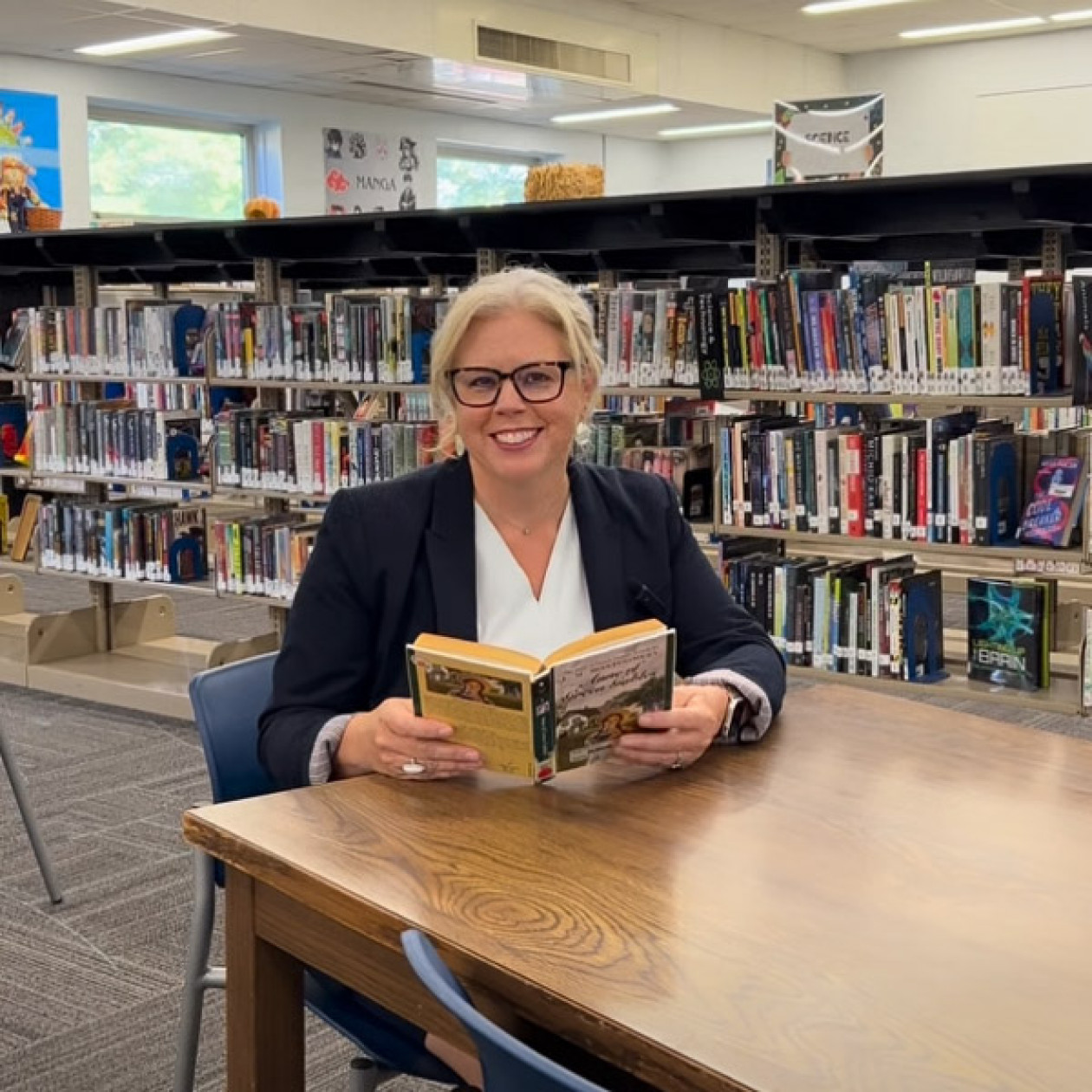 Dr. Reed sits at a desk in a media center while holding a book.