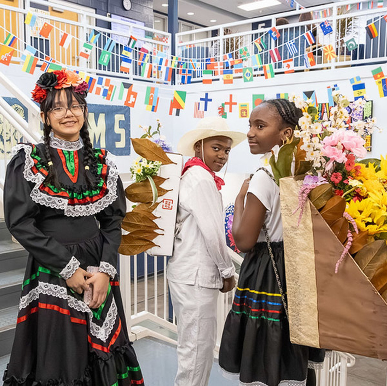 Three students pose in front of many world flags while wearing traditional clothing.