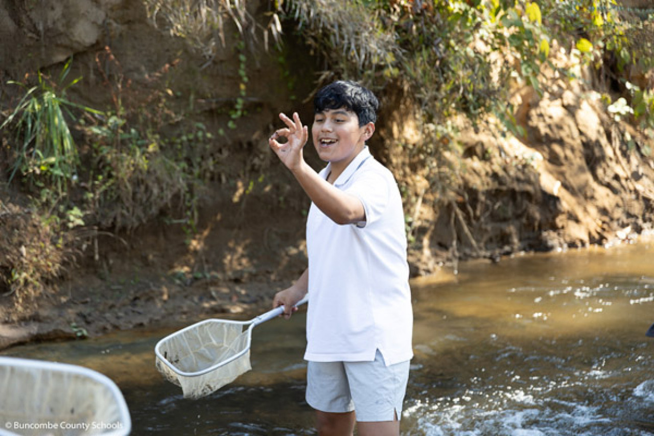 A boy holds a net in one hand and a tiny specimen in the other while standing in a creek.
