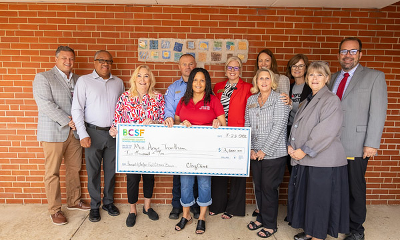 Ms. Trantham holds a giant novelty check while surrounded by family, school and district leadership