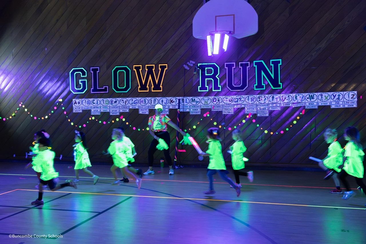 Kids in glowing neon shirts run by a sign that reads 