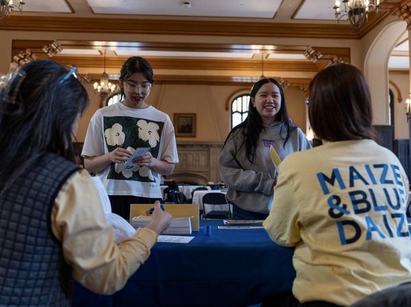 Two maize and blue daize staff sit at a table talking to two students standing on the other side of the table