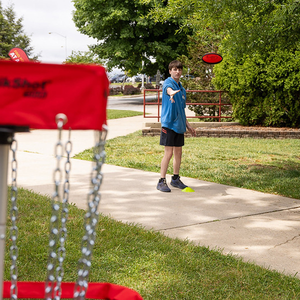 A student throws a disc at a disc golf pin.