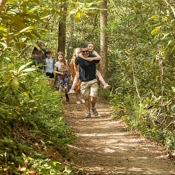 A man carries a girl on his back while a group of students walk behind on a trail.