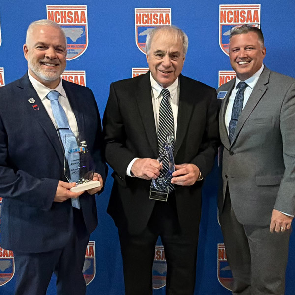 From left, Brian Carver, David Ball, and Chip Cody pose in front of an NCHSAA backdrop while Carver and Ball hold plaques.