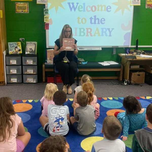 A teacher holds up a book as children sit on a rug in a classroom.