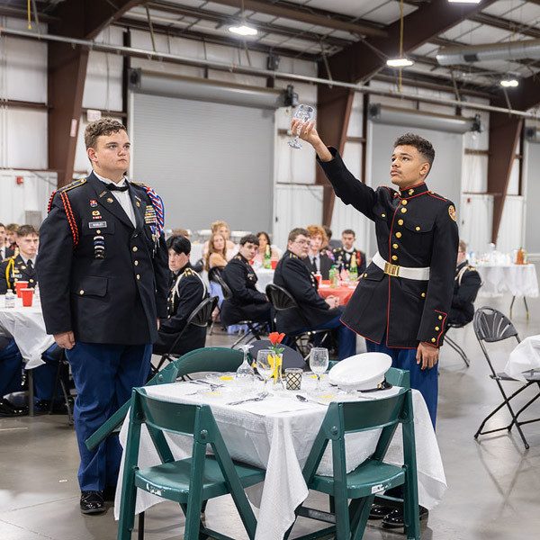 Two students, decorated in military attire, stand at a table. One stands at attention while the other lifts a glass into the air.