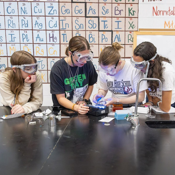 Four students, wearing safety goggles, lean over an experiment in a science lab.